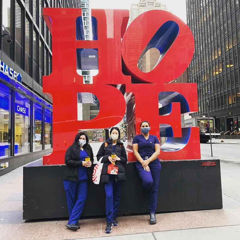 Three nurses in scrubs outside the red HOPE sculpture in midtown Manhattan