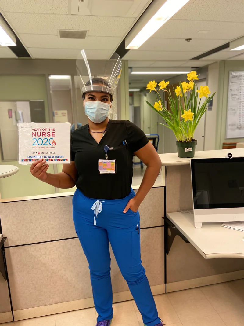 Joanna in a face shield holding a Year of the Nurse 2020 sign at the nursing station